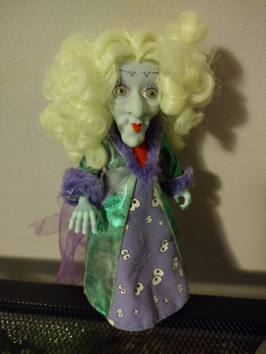 A photo of a doll with bright green, curly hair, light green skin, crazy green eyes, and bright red lipstick. She's wearing a long, shimmery, green gown with purple cuffs. The gown has spooky eyes on the front.